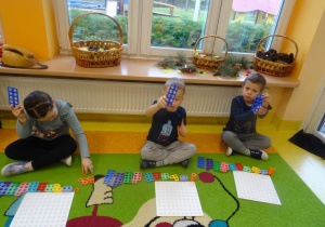 Troje dzieci siedzi na dywanie z ułożonymi przed nimi płytkami i klockami Numicon, w uniesionych dłoniach trzymają klocek z 10 otworami.